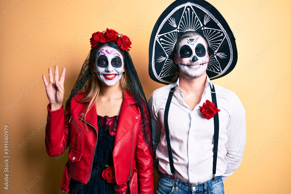 Couple wearing day of the dead costume over yellow showing and pointing up with fingers number four while smiling confident and happy.