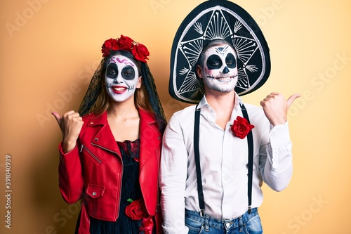 Couple wearing day of the dead costume over yellow smiling with happy face looking and pointing to the side with thumb up.