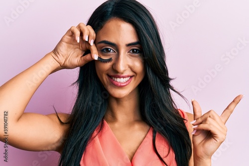 Beautiful hispanic woman holding fake lashes smiling happy pointing with hand and finger to the side