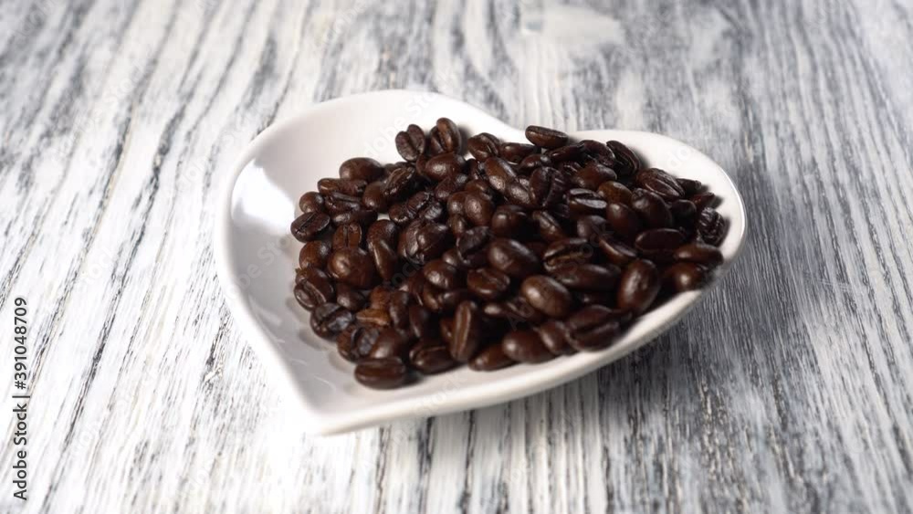 Roasted coffee beans are poured from hands into a white saucer. Heart shape. On a wooden background close up. Valentine Day Breakfast Roasted coffee beans are poured from hands into a white saucer. Heart shape. On a wooden background close up. Valentine Day Breakfast