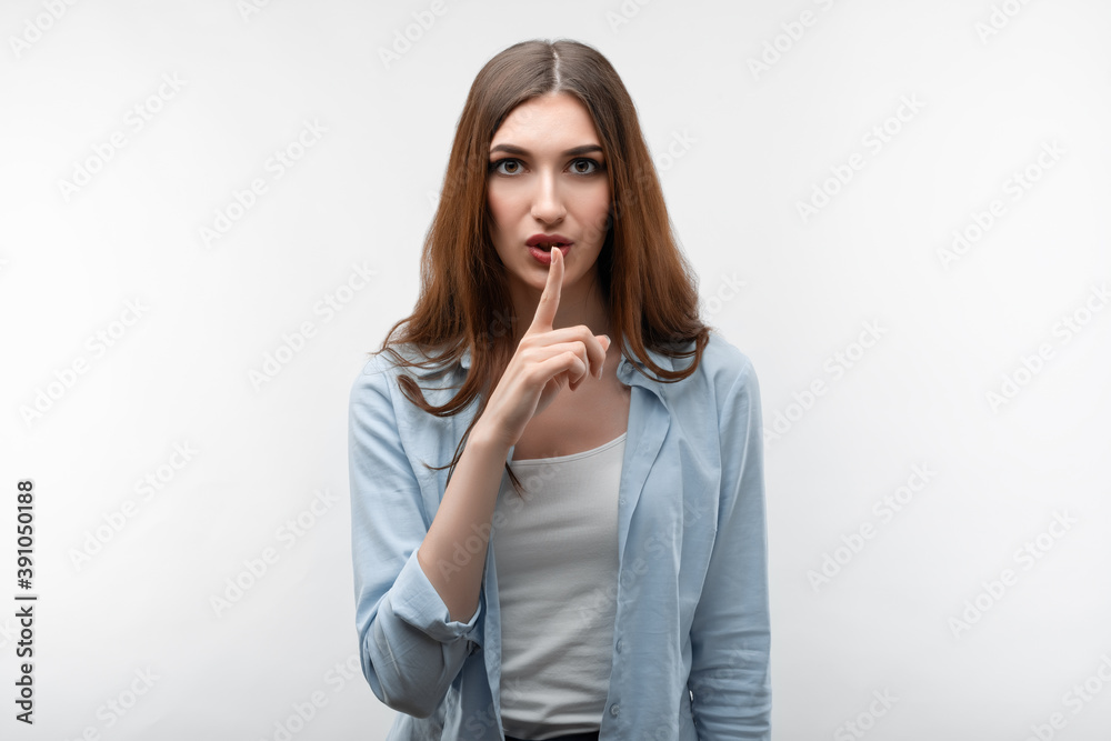 Pretty girl keeps finger on lips, making hush gesture on white background. Shh, silence concept