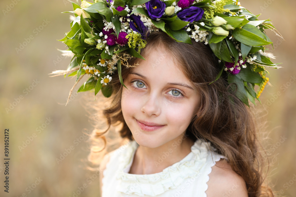 Cute little girl wearing wreath made of beautiful flowers in field on sunny day Cute little girl wearing wreath made of beautiful flowers in field on sunny day