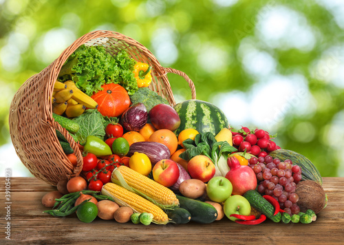 Fototapeta Naklejka Na Ścianę i Meble -  Overturned wicker basket with different fresh organic vegetables and fruits on wooden table against blurred green background