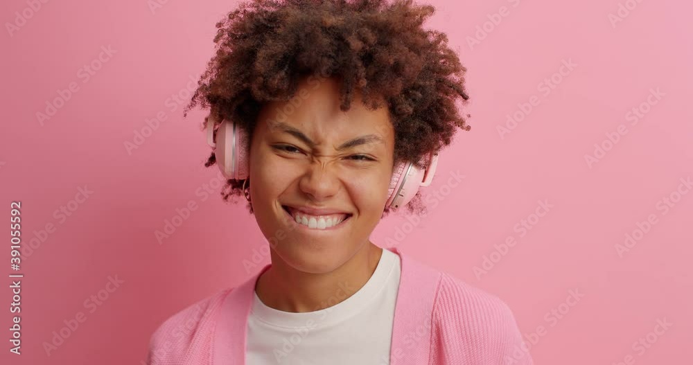 Joyous dark skinned woman smiles broadly sends positive vibes listens music in stereo headphones moves carefree poses against pink background enjoys great vibes isolated over pink background Joyous dark skinned woman smiles broadly sends positive vibes listens music in stereo headphones moves carefree poses against pink background enjoys great vibes isolated over pink background