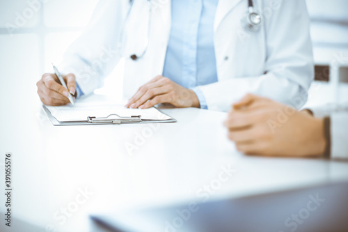 Unknown woman-doctor and female patient sitting and talking at medical examination in clinic  close-up. Therapist wearing blue blouse is filling up medication history record. Medicine concept