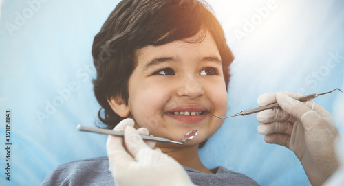 Cute arab boy sitting at dental chair with open mouth during oral checking up with doctor. Visiting sunny dentist office