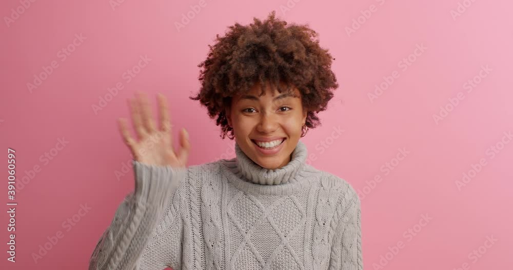 Cheerful friendly dark skinned millennial girl says hello and waves hand to welcome friends smiles broadly shows hi gesture greets someone with happy expression dressed casually poses indoor Cheerful friendly dark skinned millennial girl says hello and waves hand to welcome friends smiles broadly shows hi gesture greets someone with happy expression dressed casually poses indoor