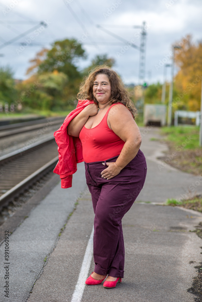 Sehr mollige Frau mittleren Alters in Top und Jeans in lässiger Pose mit Lederjacke wartet auf den Zug am Bahnsteig