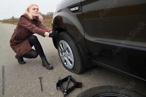 A young blonde woman removes the wheel with a key near her car with a flat tire.