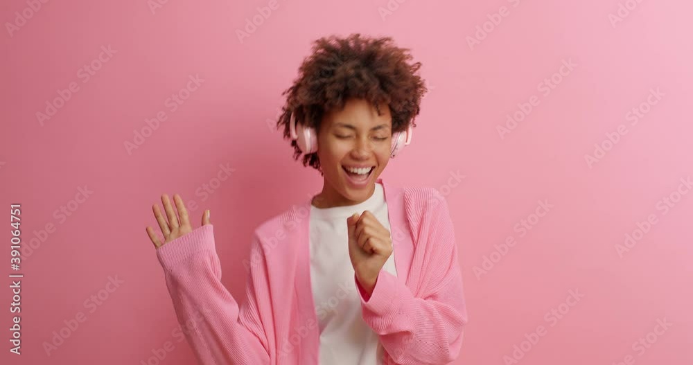 Overjoyed dark skinned ethnic woman moves with rhythm of music wears headphones and sings favorite song dressed in casual clothing isolated over pink background. Slow motion. Carefree dance. Overjoyed dark skinned ethnic woman moves with rhythm of music wears headphones and sings favorite song dressed in casual clothing isolated over pink background. Slow motion. Carefree dance.