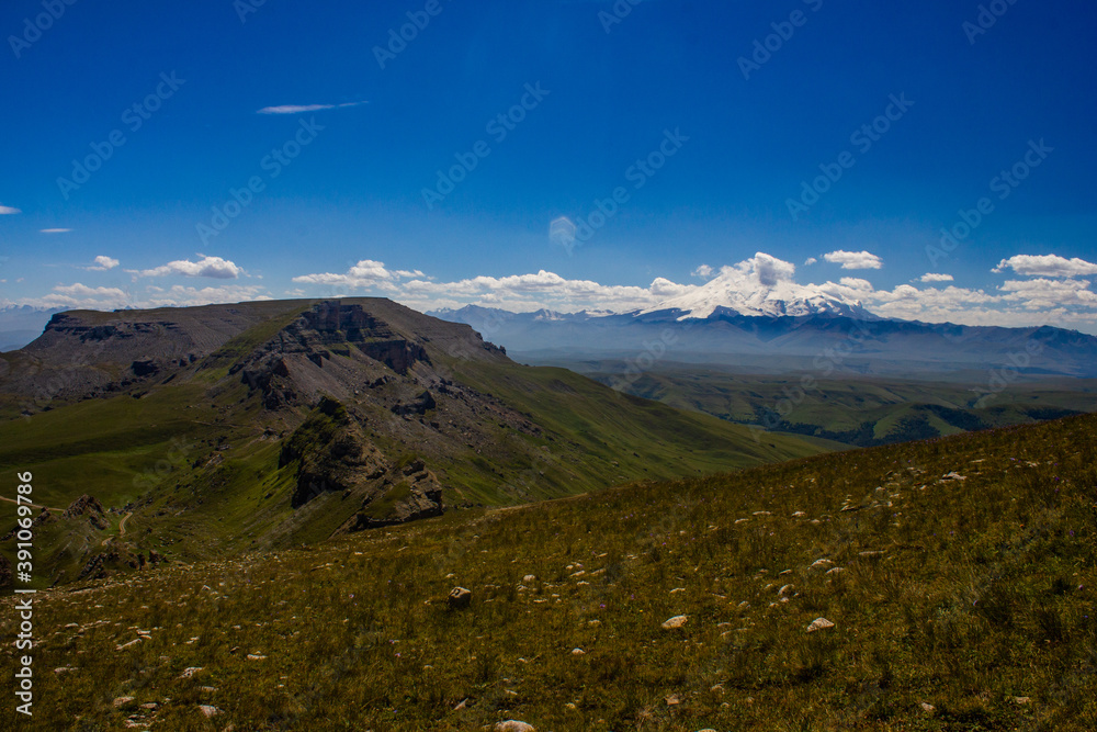 An excellent view from the plateau to the extinct volcano and the snowy peak of Mount Elbrus. Below is a green gorge An excellent view from the plateau to the extinct volcano and the snowy peak of Mount Elbrus. Below is a green gorge