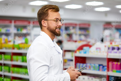 side view on handsome young pharmacist man using smartphone at work, texting messaging consulting client or patient on cell online, browsing Internet on smartphone