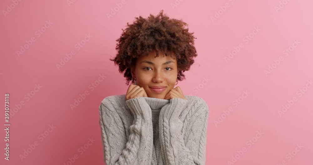 Happy satisfied African American woman with tender expression wears comfortable warm sweater keeps hands on collar feels calm and pleased looks tenderly at camera isolated over pink background. Happy satisfied African American woman with tender expression wears comfortable warm sweater keeps hands on collar feels calm and pleased looks tenderly at camera isolated over pink background.