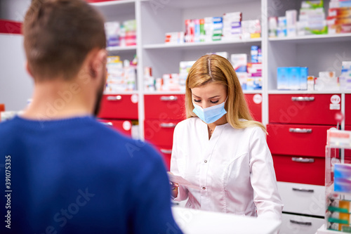young female apothecary in protective medical mask and young caucasian man customer buying drug at drugstore. medicine  consumerism concept. during coronavirus epidemic