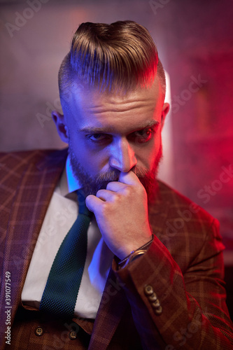 young caucasian handsome bearded man in suit sitting on couch in a dark smoky room, he is elegant and attractive, male is lost in thought. dramatic evening, dark shadows and wealth