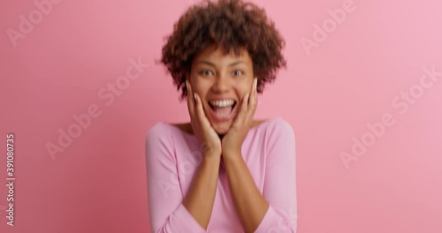 Beautiful greatly surprised woman stares at camera in amazement with bugged eyes and widely opened mouth has very happy reaction on incredible news jumps from joy poses against pink background. photo
