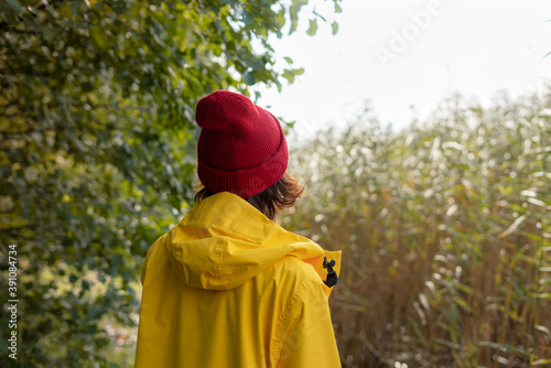 Back view of woman in bright yellow raincoat and red hat looks at reeds, unity with nature. Cold autumn day, sunshine on bay, one with nature