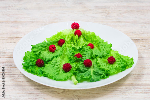 The Christmas tree is lined with green lettuce leaves, decorated with red raspberries on a white plate. Food for the New Year. Table decoration.