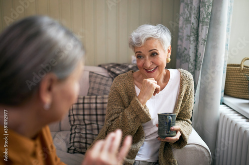 Two elderly female friendly drinking coffee together, sharing news, having cheerful facial expression. Retired senior woman with short gray hair enjoying coffee during conversation with her neighbor