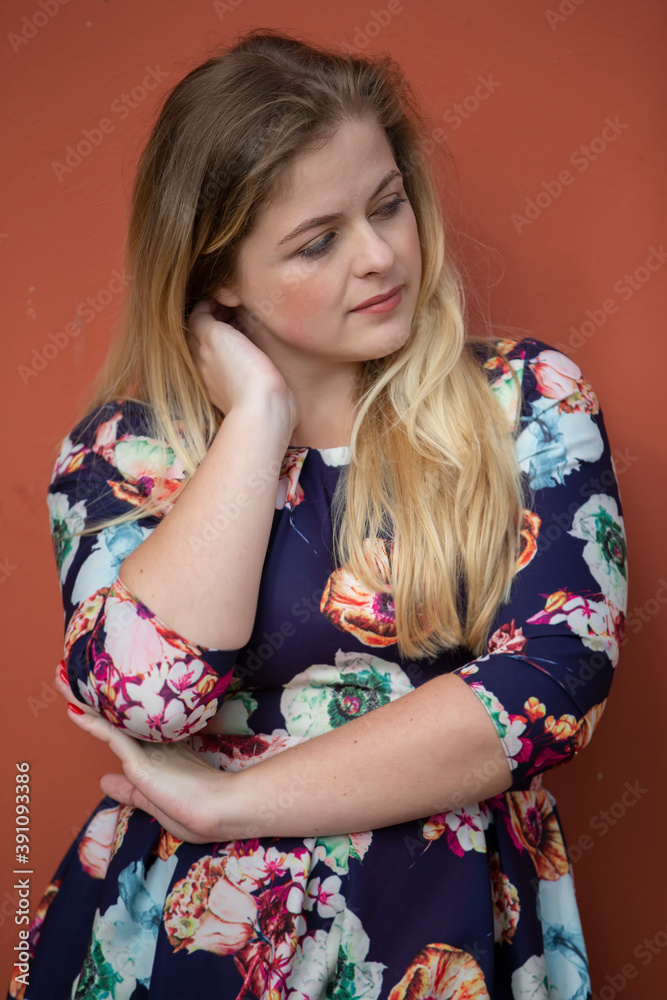 A pensive blonde girl with blue eyes in a blue dress with a floral print stands near a burgundy wall. A pensive blonde girl with blue eyes in a blue dress with a floral print stands near a burgundy wall.