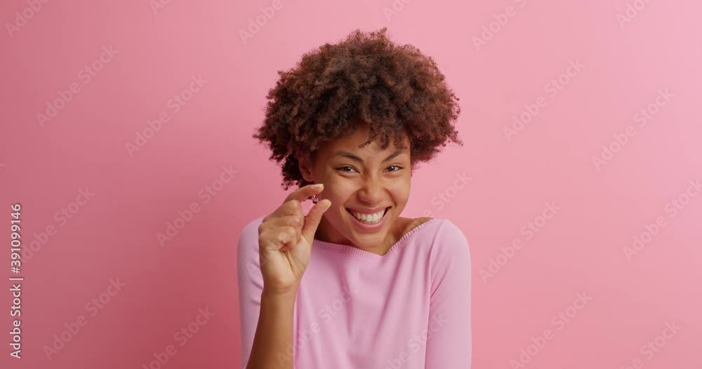 Young brunette young woman with curly Afro hair shows very tiny object shapes small item needs some more dressed in casual clothing poses against pink background. Minimum size. I need not much Young brunette young woman with curly Afro hair shows very tiny object shapes small item needs some more dressed in casual clothing poses against pink background. Minimum size. I need not much