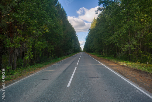 An asphalt straight road in the middle of the forest against a background of blue sky and white clouds.