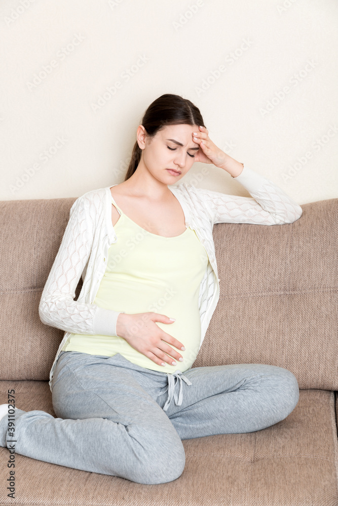Young pregnant woman with headache sitting on sofa. Pregnancy expectation concept Young pregnant woman with headache sitting on sofa. Pregnancy expectation concept