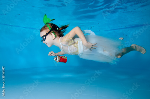 Portrait of a beautiful little girl with a Christmas toy in her hand and a white dress. She swims underwater in a pool on a blue background and smiles. Horizontal orientation