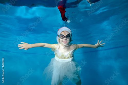 Portrait of a cheerful little girl in a Santa hat and white dress, who swims and poses for the camera underwater in a children's pool and smiles. Concept. Horizontal orientation
