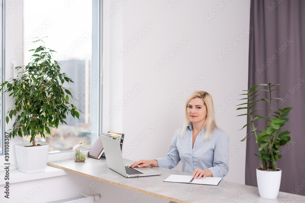 Young blonde caucasian woman photographer sitting at the laptop wooden table large window and grey wall working Young blonde caucasian woman photographer sitting at the laptop wooden table large window and grey wall working