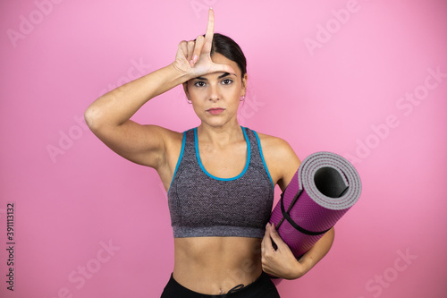 Young beautiful woman wearing sportswear over isolated pink background holding a splinter in her hand making fun of people with fingers on forehead doing loser gesture mocking and insulting.