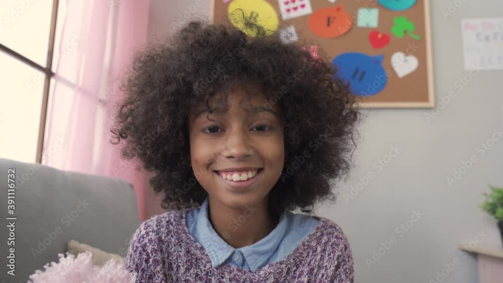 Happy cute small african kid girl vlogger talking to camera at home recording vlog for social media blog, video conference calling virtual friend having online meeting sitting on couch, web cam view.