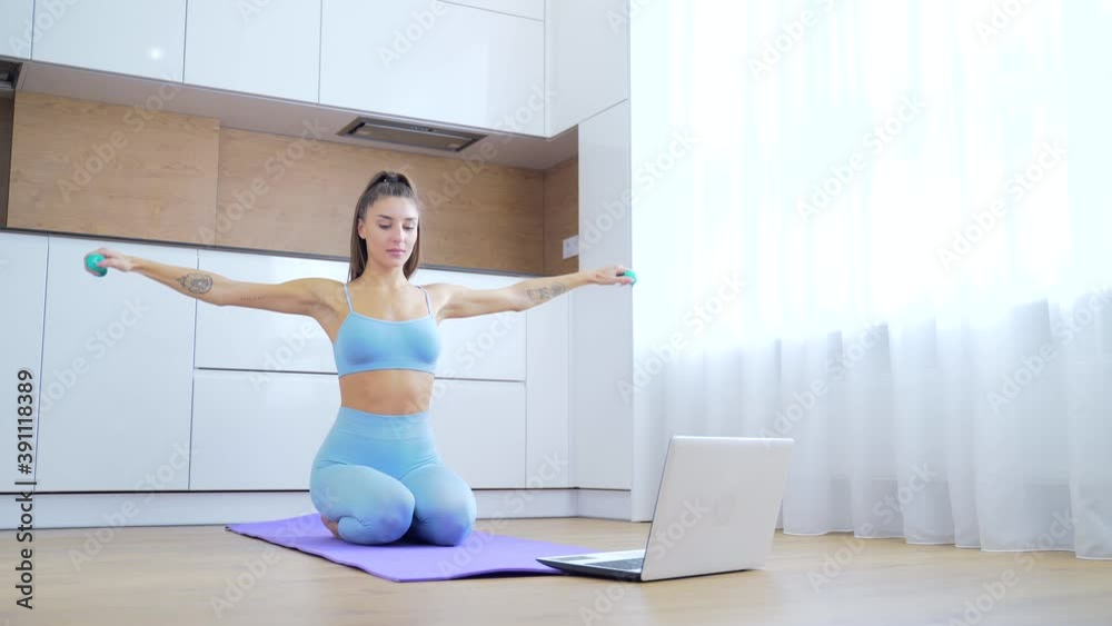young attractive fitness woman in sportswear exercising online with dumbbells in front of laptop in kitchen sitting on the floor. girl goes sports at a distance on lessons training quarantine at home