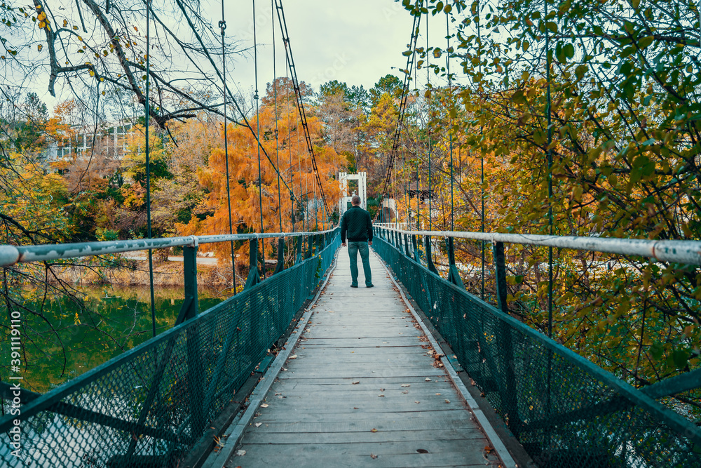 Obraz premium Old suspension bridge over river and man at gray autumn day. Melancholy, depression or loneliness.
