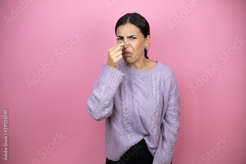 Beautiful woman wearing a casual violet sweater over pink background smelling something stinky and disgusting, intolerable smell, holding breath with fingers on nose © Irene