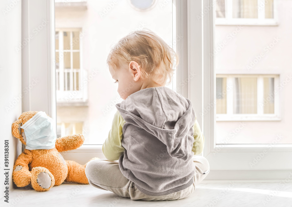 Child in home quarantine standing at the window with his sick teddy bear wearing a medical mask against viruses during coronavirus and flu lock down. Children and illness COVID-2019 disease concept Child in home quarantine standing at the window with his sick teddy bear wearing a medical mask against viruses during coronavirus and flu lock down. Children and illness COVID-2019 disease concept