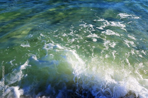 The green wave comes from the ocean, breaks into splashes on the shore and foams, the water is clear, the bottom is visible through it, close-up
