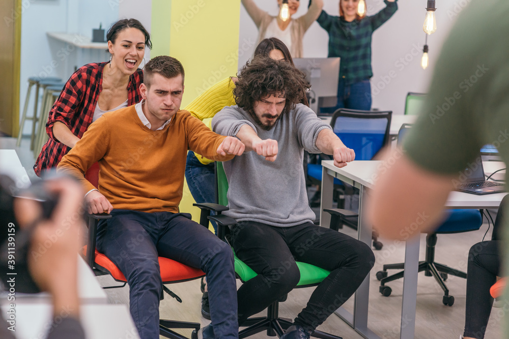 Obraz premium Team building and office fun.Four young cheerful businesspeople in smart casual wear having fun while racing on office chairs and smiling..