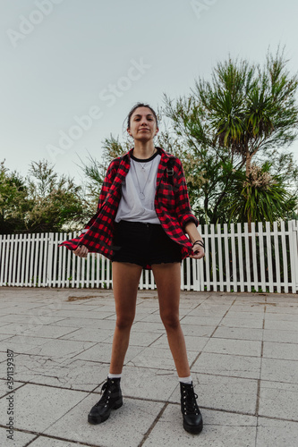 Young beautiful brunette woman in casual plaid red shirt standing, trees background, outdoor in the city, fashion concept, happy woman, copy space, social media, urban style © Rodrigo Rivas 