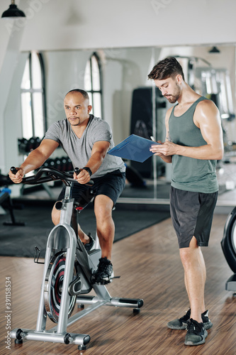 Concentrated mature man riding on stationary bike when trainer writing results in document