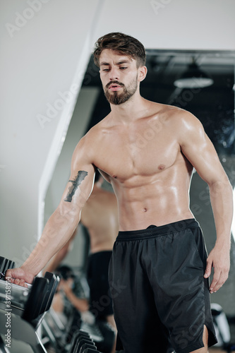 Sweaty young fit strong man taking heavy dumbbell from stand in gym