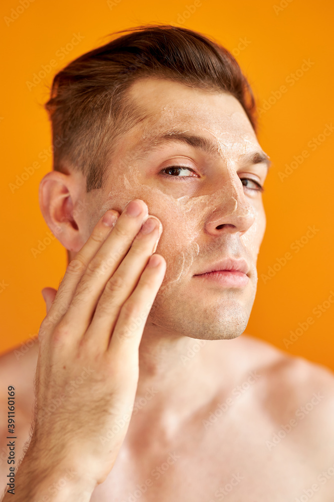 Obraz premium good-looking caucasian man cares for his skin, moisturizes it with professional cosmetics product, isolated orange background