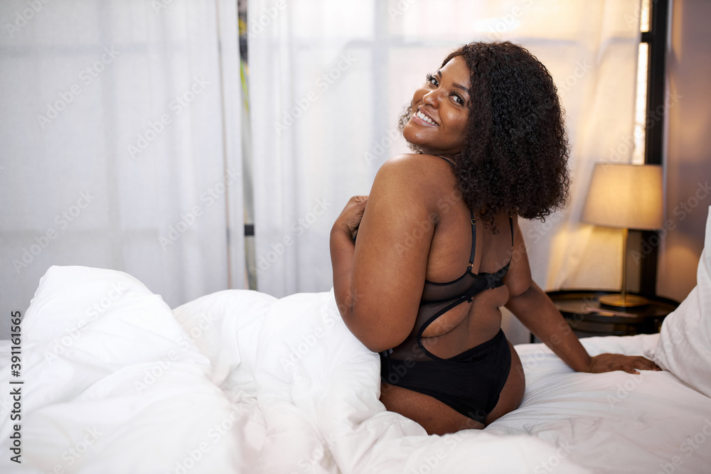 adorable afro woman smile sitting on bed, has happy day, enjoy being fat, she has no complexes about her appearance adorable afro woman smile sitting on bed, has happy day, enjoy being fat, she has no complexes about her appearance