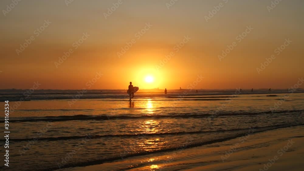 A beautiful young surfer in silhouette carries his surfboard down into the shallow water for a sunset surf session on a tropical beach. Slow motion cinematic shot. A beautiful young surfer in silhouette carries his surfboard down into the shallow water for a sunset surf session on a tropical beach. Slow motion cinematic shot.