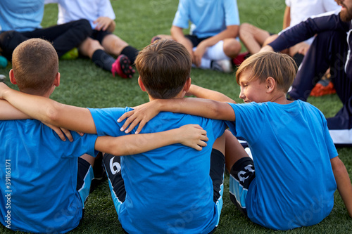 young kids football team embrace each other on football field before match, support, have talk, sit on grass together © Roman