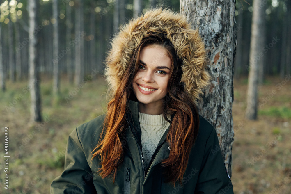 Woman in warm jacket with a hood near a tree in the forest smile attractive look Woman in warm jacket with a hood near a tree in the forest smile attractive look