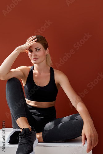 attractive woman with pepfect body sits on box relaxing isolated over red background, young female in black sportswear