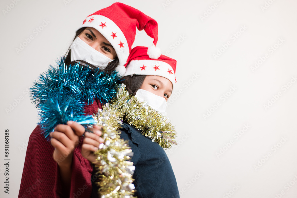 Mother and daughter looking to the camera wearing santa hat and surgical mask holding tinsel sparkled on white background with copy space for text.