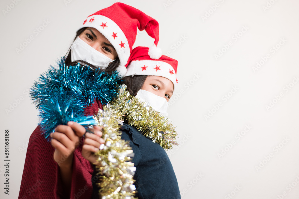 Mother and daughter looking to the camera wearing santa hat and surgical mask holding tinsel sparkled on white background with copy space for text. Mother and daughter looking to the camera wearing santa hat and surgical mask holding tinsel sparkled on white background with copy space for text.