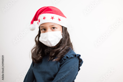 Little girl wearing santa hat and face mask looking to the camera sad on white background.